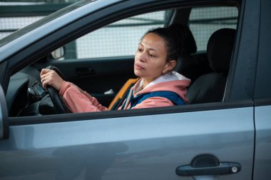 Mavi bir arabanın direksiyonunda oturan bir kadın, pembe kapüşonlu ve mavi ceketli, gündelik bir sürüş sırasında dalgın dalgın görünüyor. İnce, her gün.