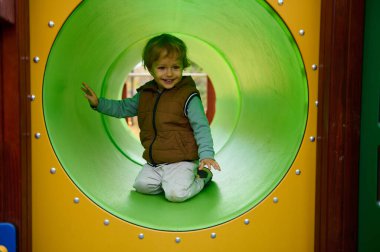 Kahverengi yelek giyen neşeli bir çocuk yeşil bir oyun parkı tünelinde diz çöküp oynarken gülümsüyor. Kaygısız çocukluk, açık hava eğlencesi ve eğlenceli keşifleri yakalar..