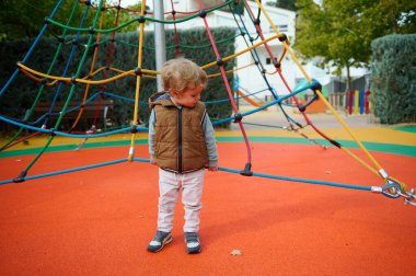 Kahverengi yelekli bir çocuk, oyun parkında canlı bir halat tırmanma yapısını keşfediyor, neşeli bir sonbahar gününün ve sıcak bir aile ortamının keyfini çıkarıyor..