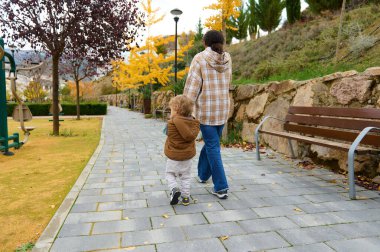 Bir anne ve çocuk sonbahar yaprakları arasında asfalt bir park yolunda yürürler, nazik bir bağ ve sıcak, kaygısız bir anın tadını çıkarırlar..