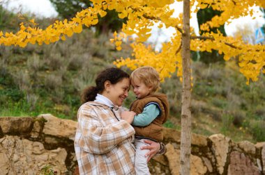 Sevgi dolu bir anne bebeğini bir parkta tutar, altın sonbahar yaprakları arasında gülümser ve taş bir duvar zemininde, bakım, neşe ve aile birlikteliğini taşır..