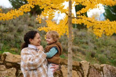 Sevgi dolu bir anne, bebeğini açık havada, parlak sonbahar yapraklarının arasında sıcacık bir gülümsemeyle tutar. Bir park ortamında aile, bakım ve mutluluk anı..