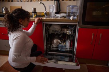 Bir kadın açık bir bulaşık makinesinin yanında diz çökmüş, açık kırmızı bir mutfakta bulaşıkları yıkıyor. Sahne, günlük ev yaşamını, ev işlerini ve aile rutinini sıcak aile ortamında yakalıyor..