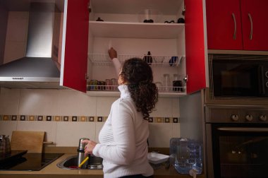 Beyaz kazaklı bir kadın açık kırmızı mutfak dolaplarına uzanıyor, modern bir ev mutfağında paslanmaz aletlerle yemek hazırlarken bardakları ve tabakları düzenliyor..