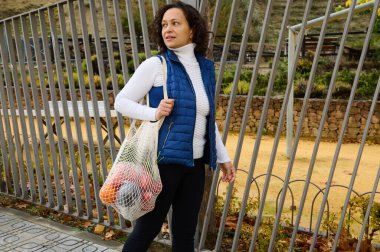 Bir kadın, alışveriş torbalarıyla dolu kullanılabilir bir ağ torbası taşıyarak park yolunda yürüyor. Beyaz balıkçı yaka ve mavi kabarık yelek giyiyor. Arka planda metal çitli ve doğal manzaralı..