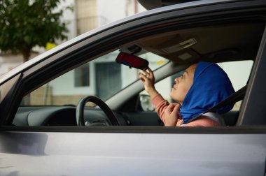 Mavi başörtüsü takan Müslüman bir Arap kadın, bir arabada oturur ve modern günlük hayatı ve bağımsızlığı sembolize ederek, Avrupa 'nın kentsel ortamında araba sürerken seyreder..