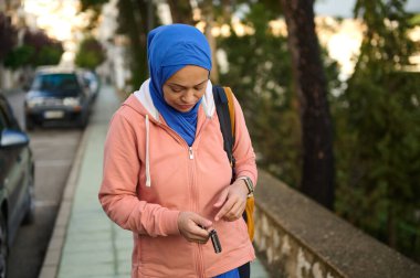 Mavi başörtüsü ve şeftali rengi kapüşonlu Müslüman bir kadın şehir kaldırımında duruyor, modern Avrupa 'da yoğun bir gün için anahtarlarını kontrol ediyor..