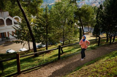 Türbanlı bir kadın, ağaçlar, ahşap çitler ve uzak bir şehir ile güneşli bir tepe boyunca koşar. Modern, sağlıklı bir açık hava yaşam tarzı..