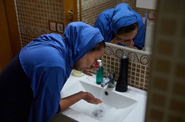 Mavi tesettürlü Müslüman bir Arap kadın modern bir Avrupa evindeki banyo lavabosunda yüzünü yıkıyor. Geleneği çağdaş yaşamla harmanlayan bir geleneği resmediyor..