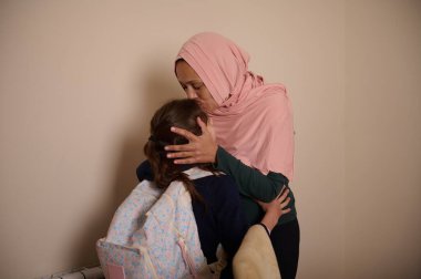 Pembe tesettürlü Müslüman bir anne, çocuğunu evde nazik bir öpücükle rahatlatır. Huzurlu bir yuvada sıcaklığı, ilgiyi ve yakın aile bağını aktarır..