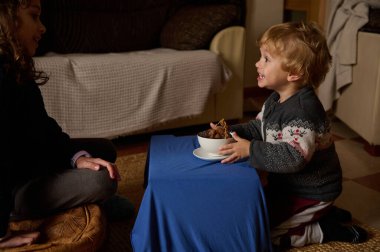 Neşeli bir süveter giyen gülümseyen bir çocuk sıcak aydınlatılmış bir oturma odasında bir arkadaşla bir kase ikramı paylaşıyor ve sağlıklı bir aile ve dostluk anı yakalıyor..