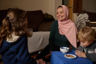 Pembe başörtüsü takan Müslüman bir Arap kadın, arkadaşlarıyla ve çocuğuyla birlikte alçak bir masada oturur. Sıcak bir oturma odasında atıştırmalık ve içecek paylaşırken gülümserler..