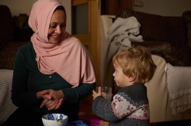 Pembe tesettürlü Müslüman bir Arap kadın yürümeye başlayan çocuğuna gülümserken evlerinde sakin ve sıcak bir an paylaşıyorlar, günlük aile yaşantılarını ve bağlantılarını yansıtıyorlar..