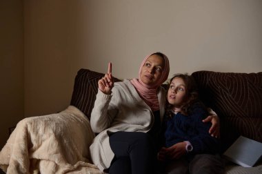 Müslüman bir Arap kadın tesettürde kızıyla birlikte rahat bir kanepede oturur. Parlak bir oturma odasında sıcak ve samimi bir anı paylaşırken yukarıya doğru işaret eder..