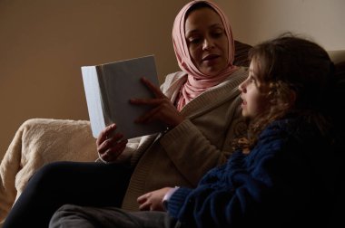 Başörtülü Müslüman bir kadın kanepeye oturur ve genç bir kıza kitap okur. Sıcak, samimi bir ev sahnesi aile hayatını ve öğrenmeyi modern bir ortamda gösteriyor..