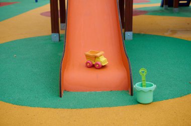 Turuncu kaydırağlı canlı bir oyun alanı sahnesi, sarı oyuncak damperli bir kamyon, ve çok renkli kauçuk yüzeyinde küreği olan yeşil bir kum kovası, açık hava aktivitesini ve çocukluk hayalgücünü yakalıyor..