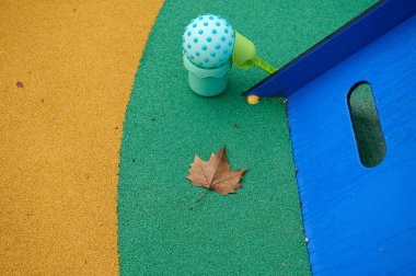 Yeşil çimenlikli renkli oyun alanı, sarı zemin, mavi kaydırak, ve kahverengi sonbahar kolyesi, açık hava eğlencesi ve mevsimsel değişiklikleri çağrıştıran eğlenceli bir ortam..