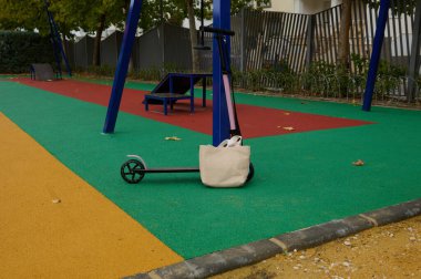 Küçük bir scooter, canlı, çok renkli bir yüzey üzerinde mavi oyun alanı ekipmanlarına karşı duruyor. Yanında bej bir çanta duruyor. Bu da parkta ya da dışarıda boş bir gün olduğunu gösteriyor..