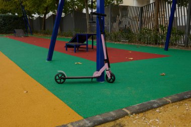Pembe bir scooter, sakin bir park ortamında parlak renkli bir lastik yüzeyde mavi bir oyun parkına yaslanır..