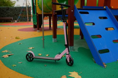 Pembe bir scooter, güneşli bir parktaki mavi bir kaydırağın yanında canlı bir oyun parkında dinlenir. Sonbaharı andıran yerde yapraklar, neşeli, aile dostu bir açık hava sahnesi yaratır..