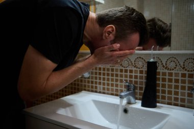 Siyah gömlekli bir adam, aynada yansıyan banyo lavabosunda yüzünü yıkıyor. Döşeme duvarı, sabun makinesi ve modern musluk sakin ve arındırıcı bir kişisel bakım anı yaratıyor..