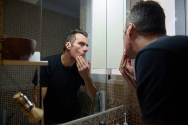 Bir adam banyo lavabosunun önünde dikilip aynada kendine bakarken cilt bakımı yapıyor. Yaşam tarzı ve kişisel bakım temaları için uygun sakin, özel bir bakım anı.