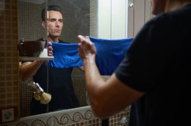 Bir adam elinde mavi bir havluyla banyo aynasında duruyor ve güne hazırlanıyor. Uzaktan çalışan bir baba ve girişimci olarak rolünü yansıtıyor..