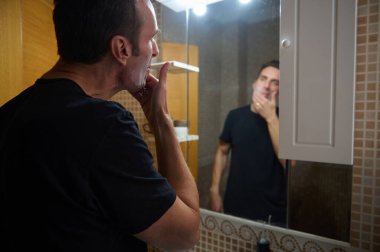 Siyah tişörtlü bir adam banyo aynasında tıraş olur, sabah rutinini yakalar ve evden gelen uzaktan kumandalı işlere odaklanır..