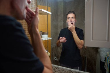 Siyah tişörtlü bir adam, banyo aynasında görülen diş macunu ve köpük sürüyor. Bu gizli çekim günlük bakım, kişisel bakım ve evde sakin bir sabah rutinini yakalar..