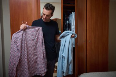 Bir adam yatak odasındaki dolapta duruyor, bir elinde kareli bir gömlek, diğer elinde açık mavi bir kapüşonlu, evde sıradan bir gün için ne giyeceğini seçiyor..