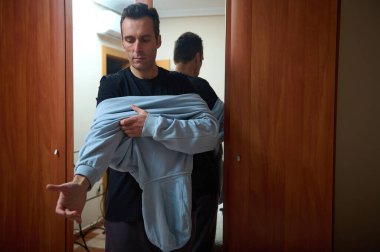 Bir adam ahşap bir kapıda dikiliyor, koyu renk bir gömlek giyerken mavi bir kapüşonluyu kaldırıyor, düşünceli görünüyor. Ev hayatı, aile, iş ve günlük anlar için uygun..