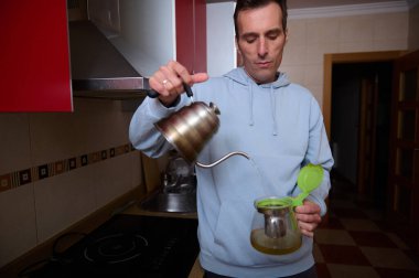 Açık mavi kapüşonlu bir adam evdeki cam bir kahve sürahisine sıcak su dökerek modern bir mutfakta sabah içkisine başlıyor..