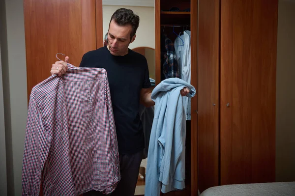 Bir adam yatak odasındaki dolapta duruyor, bir elinde kareli bir gömlek, diğer elinde açık mavi bir kapüşonlu, evde sıradan bir gün için ne giyeceğini seçiyor..