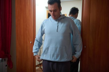 Açık mavi kapüşonlu bir adam, evdeki yatak odasındaki aynadaki uyumu kontrol ediyor, rahat ve rahat bir yaşam tarzı ve moda malzemesi kullanımı için evden işe geçen bir anı vurguluyor..