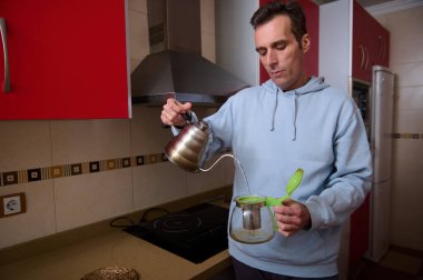 Açık mavi kapüşonlu bir adam evde çay hazırlıyor, yeşil kapaklı cam bir çaydanlığa sıcak su döküyor. Modern bir mutfakta sıradan bir ev havası..