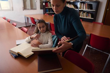 Bir öğretmen kütüphane sınıfında bir kitapla genç bir kıza yardım eder, uzun bir masada birlikte çalışırken öğrenmeyi ve odaklanmayı teşvik eder..