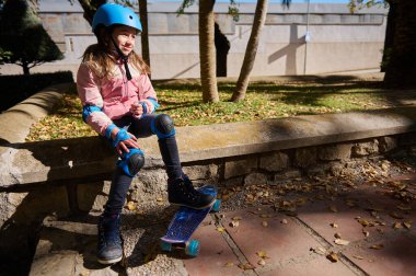 Mavi kask takan ve dizlik takan genç bir kız kaykaylı kaya çıkıntısında oturur ve güneşli bir sonbahar gününün tadını çıkarır..