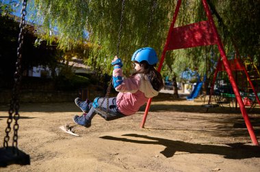 Mavi miğfer takan bir kız, güneşli bir parkta neşeli bir salıncak sürüşünden, hareketten, güvenlik donanımından ve açık hava oyununun kaygısız ruhundan zevk alır..