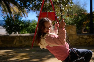 Gülümseyen genç bir kız güneşli bir parkta salıncaktan hoşlanıyor, çocukluk anlarını ve açık hava eğlencesini yakalıyor..
