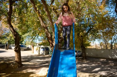 Genç bir kız parlak mavi bir oyun parkı kaydırağının tepesinde duruyor, eğlenceli bir sürüş için hazır. Etrafı sonbahar ağaçlarıyla çevrili, gülümsüyor, açık havada kaygısız bir parkı yakalıyor..