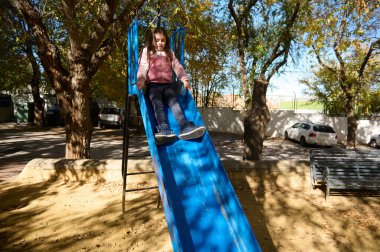 Neşeli bir kız, güneşli bir parkta parlak bir mavi kaydırağın üzerinde kayarak açık hava oyunlarını ve çocukluk sevincini yakalıyor..