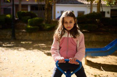 Genç bir kız, pembe ve krem rengi ceket giymiş mavi bir baharda ata biniyor. Ağaçlı güneş parkı ve arka planda bir kaydırak, kaygısız çocukluk anılarını yakalıyor..