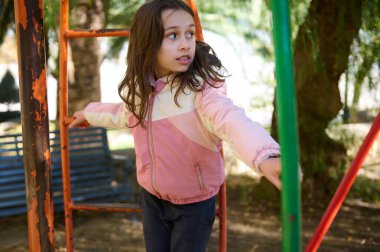 Pembe ceketli genç bir kız, elinde tuhaf bir ifadeyle metal parmaklıklara tutunarak bir oyun alanını keşfediyor. Açık hava, güneşli bir gün, oyun oynayan çocuklar, mahalle parkı..