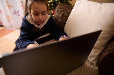 Genç bir kız dizüstü bilgisayarı ve telefonu olan rahat bir kanepede oturuyor, ekrana odaklanmış. Sıcak Noel ışıkları, arka planda oturan bir ağaç gibi parıldıyor, rahat bir ev teknolojisi anını yakalıyor..