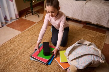 Genç bir kız örülmüş bir halıya diz çöküp renkli defterler ve sırt çantasıyla evde çalışma materyalleri hazırlıyor. Arka planda küçük bir ağaç olan rahat oda sahnesi.