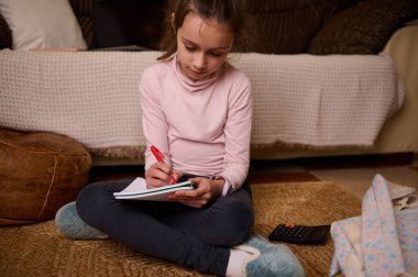 Genç bir kız örülmüş bir halıya bağdaş kurup oturma odasındaki kırmızı kalemle bir deftere yazı yazıyor. Yakınlarda bir hesap makinesi ve battaniye var..