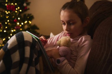 Genç bir kız kanepede oturup oyuncak ayıya sarılırken defter okuyor. Sıcak Noel ışıkları arka planda parıldıyor. Hassas, sessiz bir ev ve aile anı yaratıyor..