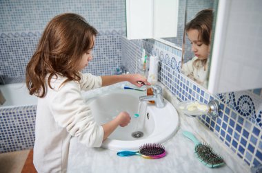 Genç bir kız banyodaki lavaboda diş fırçasını fırçalarken aynaya yansıyor. Mavi mozaik fayanslar ve bakım malzemeleri sıcak bir ev banyosunda günlük hijyen anı yaratır..