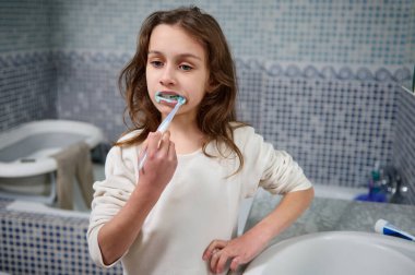 Genç bir kız banyo lavabosunda duruyor, dişlerini mavi bir diş fırçasıyla fırçalıyor. Beyaz bir gömlek giyiyor ve fayanslı duvar renkli bir zemin oluşturuyor..