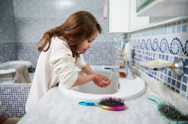Genç bir kız banyo lavabosunda ellerini yıkıyor, mavi mozaik fayanslar ve yakınlarda tımar fırçaları ile, günlük kişisel bakım ve ev anlarını yakalıyor..